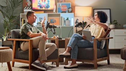 Two individuals sitting in chairs with podcasting equipment in discussion.