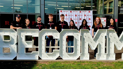 Communication majors are shown standing behind tall letters spelling out BECOMM at the Yard on the Rutgers College Avenue campus. 