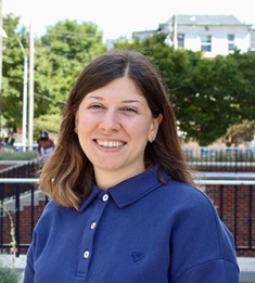 Ece Gumusel is standing outside, wearing a blue shirt, and smiling at the camera.
